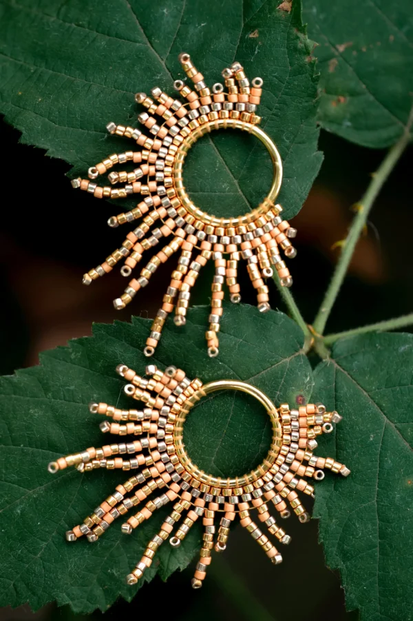 Paire de boucles d’oreilles dorées en perles Miyuki, en forme de soleil, présentées sur une feuille verte.