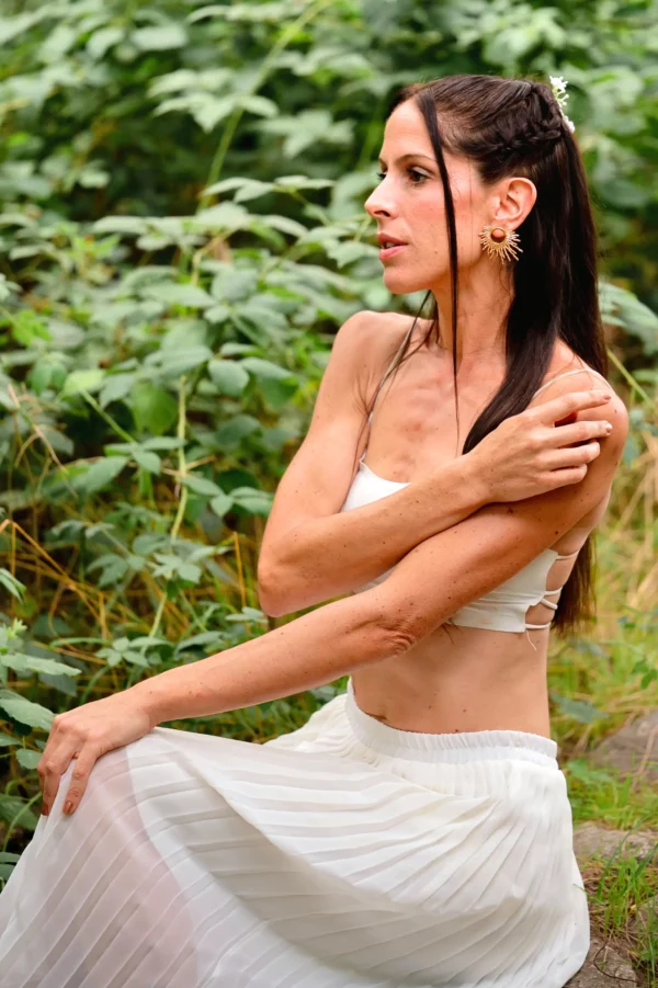 Vue latérale d’une femme habillée en blanc, assise dehors, portant les boucles d’oreilles dorées Apollisia Solaris.
