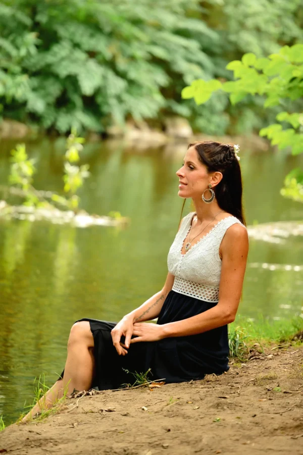 Femme assise au bord de l’eau, regardant au loin, portant les boucles d’oreilles pendantes Artemesia Lunaris Valdyria en perles Miyuki bleu et argent. Bijoux artisanaux faits main en Alsace, inspirés par la lune et la nature.