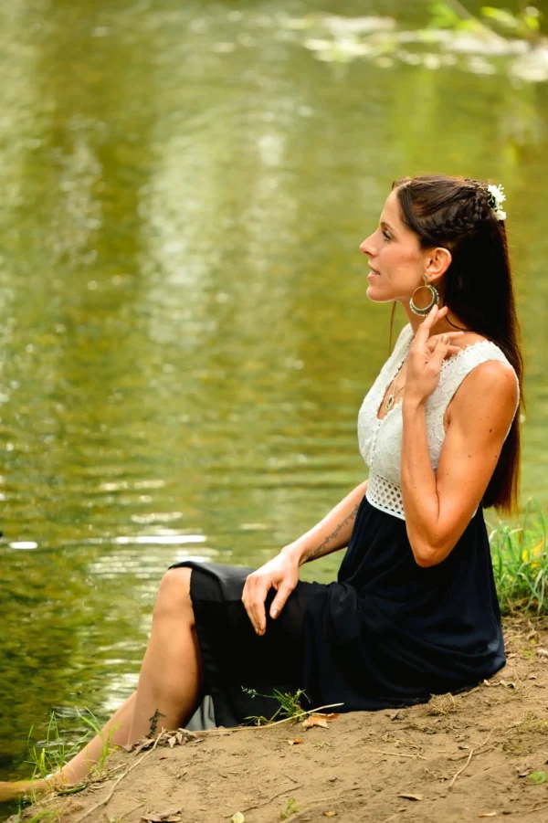 Femme en robe noire et blanche, assise près de l’eau, portant les boucles Artemesia Lunaris Valdyria. Bijou artisanal en perles Miyuki fait main.