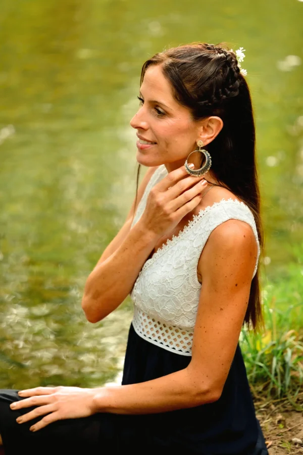 Femme de profil assise au bord d’un lac, portant les boucles Artemesia Lunaris Valdyria, bijoux artisanaux faits main en perles Miyuki bleu et argent.