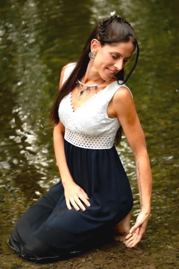 Femme asssise dans l’eau, main effleurant la surface, portant le collier Majestueux Artemesia Lunaris Valdyria en perles Miyuki noir, doré et argent. Bijou artisanal fait main.