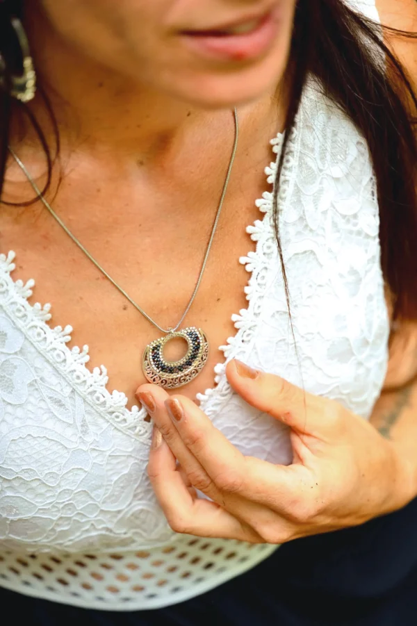 boutique-valdyria-artemesia-lunaris-collier-sautoir-gros-plan-main Femme en robe blanche tenant le pendentif du Collier Sautoir Artemesia Lunaris Valdyria sur sa poitrine. Bijou artisanal en métal ciselé et perles Miyuki.