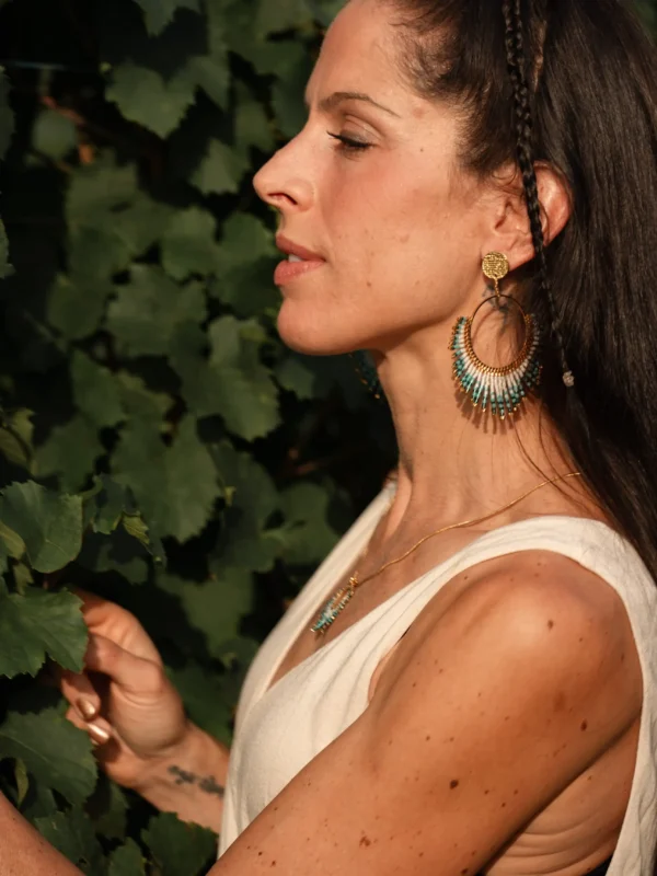 Femme de profil portant les boucles Mira Calypsa Valdyria, perles Miyuki turquoise et vert d’eau, monture dorée, regard vers l’horizon. Bijoux artisanaux français, parure inspirée des légendes d’Ulysse.