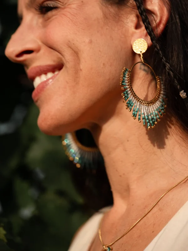 Femme souriante portant les boucles Mira Calypsa Valdyria, perles Miyuki turquoise et vert d’eau, monture dorée. Bijoux artisanaux made in France, talisman de joie.