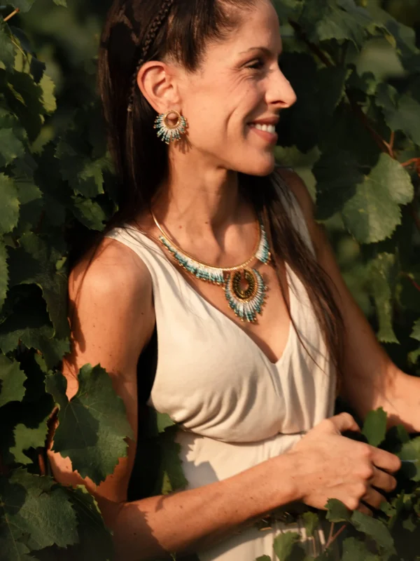 Femme souriante portant les petites boucles Mira Calypsa Valdyria et le collier assorti, perles Miyuki turquoise et blanches, monture dorée, portrait dans les vignes. Bijoux artisanaux français, slow jewelry.