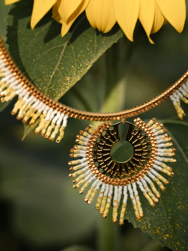 Gros plan du collier Mira Helia en perles Miyuki dorées et blanches, suspendu sur des feuilles vertes. Bijou artisanal français, confectionné à la main en Alsace, création écoresponsable et poétique.