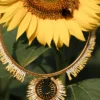 Collier majestueux Mira Helia en perles Miyuki dorées et blanches, suspendu devant un tournesol avec abeille. Bijou artisanal français, fait main en Alsace, création écoresponsable inspirée du soleil.
