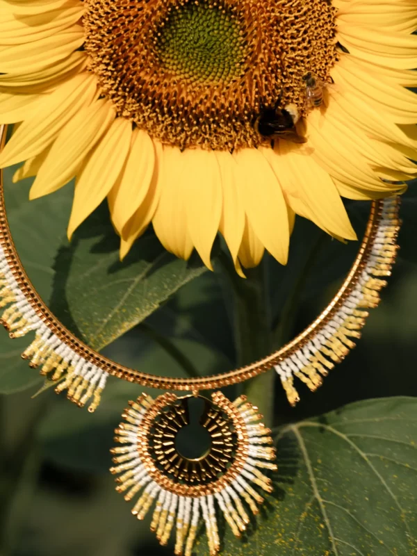 Collier majestueux Mira Helia en perles Miyuki dorées et blanches, suspendu devant un tournesol avec abeille. Bijou artisanal français, fait main en Alsace, création écoresponsable inspirée du soleil.