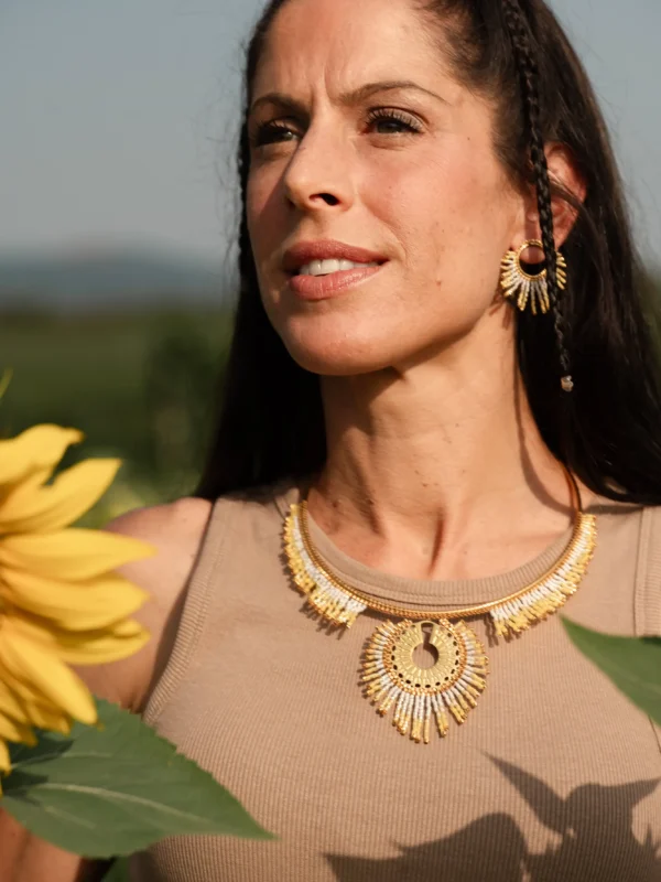 Portrait féminin avec collier majestueux Mira Helia en perles Miyuki dorées et blanches, porté dans un champ de tournesols. Bijou artisanal fait main en Alsace, création écoresponsable inspirée du soleil.