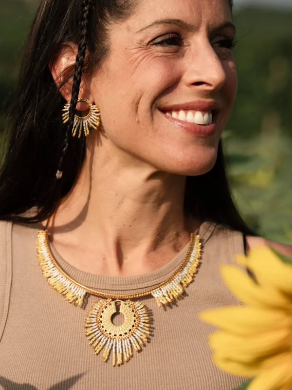 Portrait d’une femme souriante portant le collier majestueux Mira Helia et ses boucles assorties, tissés en perles Miyuki dorées et blanches. Bijou artisanal français fait main en Alsace, création durable et rayonnante.