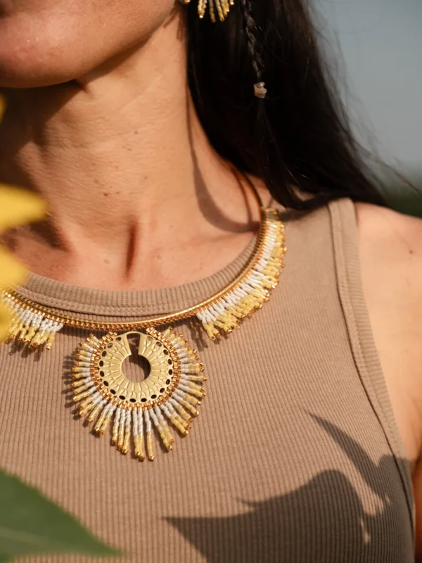 Gros plan du collier Mira Helia en perles Miyuki dorées et blanches, porté au niveau du cou, avec un tournesol en avant-plan. Bijou artisanal français, fait main en Alsace, création durable et solaire.