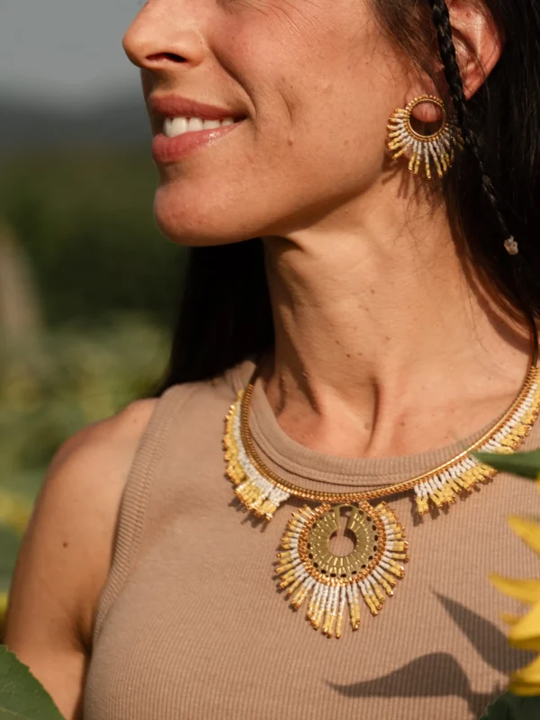 Portrait de profil d’une femme portant le collier majestueux Mira Helia et ses boucles assorties en perles Miyuki dorées et blanches. Bijou artisanal français, confectionné à la main en Alsace, création écoresponsable inspirée du soleil.