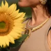 Portrait lumineux d’une femme portant le collier majestueux Mira Helia en perles Miyuki dorées et blanches, avec tournesol en avant-plan. Bijou artisanal français, fait main en Alsace, création durable et inspirée du soleil.