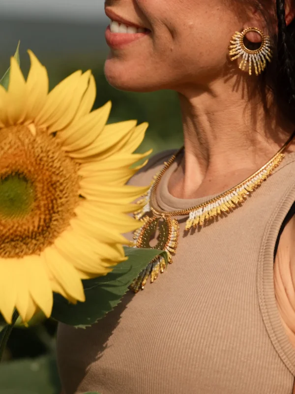 Portrait lumineux d’une femme portant le collier majestueux Mira Helia en perles Miyuki dorées et blanches, avec tournesol en avant-plan. Bijou artisanal français, fait main en Alsace, création durable et inspirée du soleil.