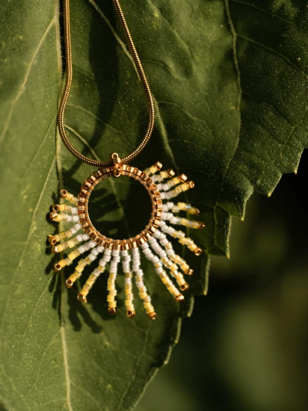 Collier sautoir Mira Helia en perles Miyuki dorées et blanches, suspendu sur une feuille illuminée. Bijou artisanal français, fait main en Alsace, création écoresponsable et solaire.