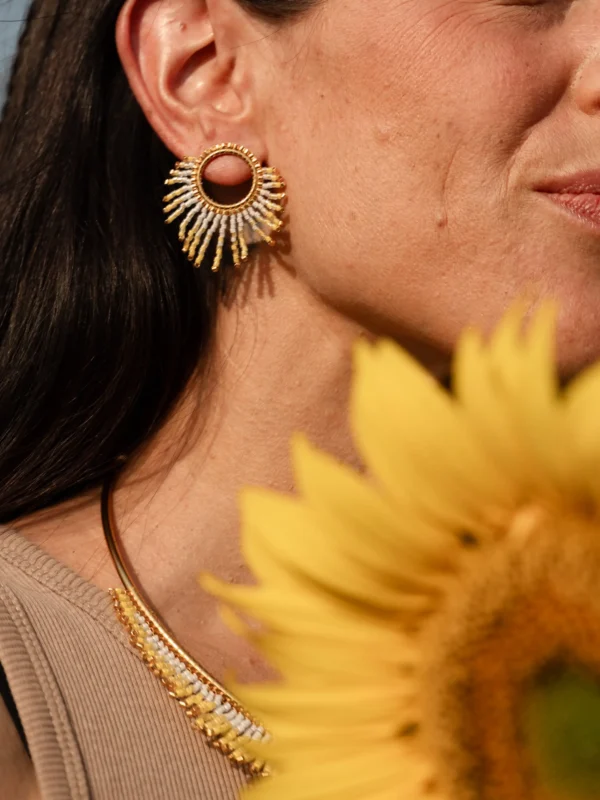 boutique-valdyria-mira-helia-petite-boucle-oreille-portrait-tournesol Petite boucle d’oreille Mira Helia en perles Miyuki dorées et blanches, portée avec un tournesol en avant-plan. Bijou artisanal français, fait main en Alsace, création durable et solaire.