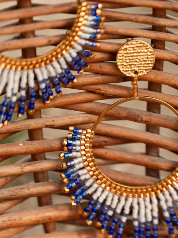 Boucle d’oreille pendante Mira Obscura en perles Miyuki bleues, blanches et dorées, posée sur fond en bois tressé. Bijou artisanal français, fait main en Alsace, création écoresponsable et poétique.