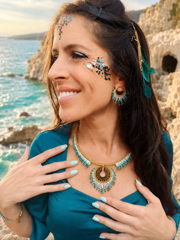 Collier majestueux Mira Calypsa porté avec main sur le décolleté, bijoux coordonnés et décor côtier naturel