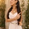 Boucles d’oreilles pendantes Mira Aurora portées en portrait avec collier assorti, robe blanche et jardin ancien lumineux