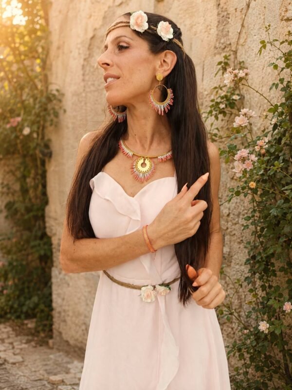 Boucles d’oreilles pendantes Mira Aurora portées en portrait avec collier assorti, robe blanche et jardin ancien lumineux