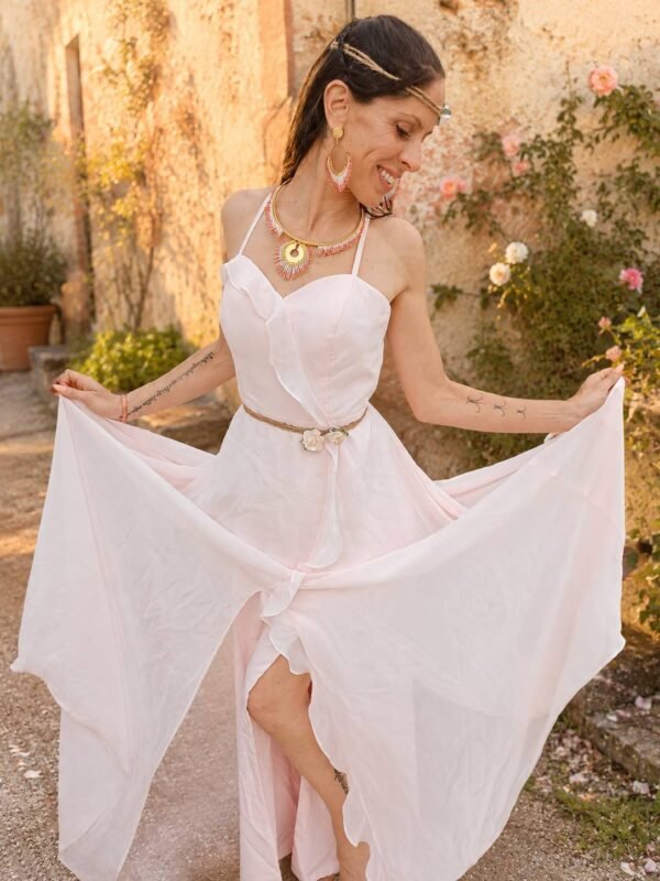 Collier majestueux Mira Aurora porté en pied avec robe claire dans un jardin fleuri, bijou fait main Valdyria aux tons rose aurore Collier majestueux Mira Aurora et nouveau départ