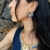 Petites boucles d’oreilles Mira Obscura portées avec collier artisanal sur robe bleu nuit dans un décor rocheux